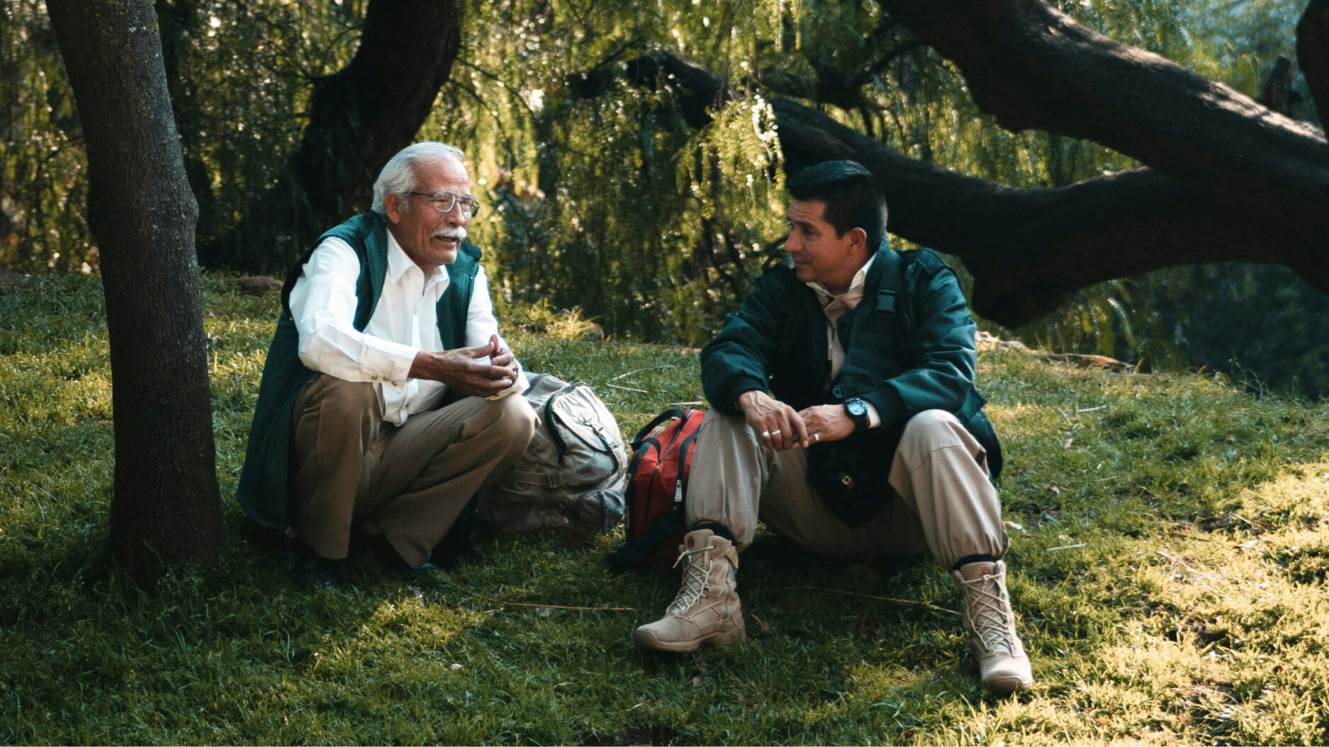 Two men sit on grass under trees, talking. One, an elderly man with white hair and a mustache, appears to be a Great Mentor. Both wear outdoor jackets; backpacks rest beside them in the dappled sunlight.