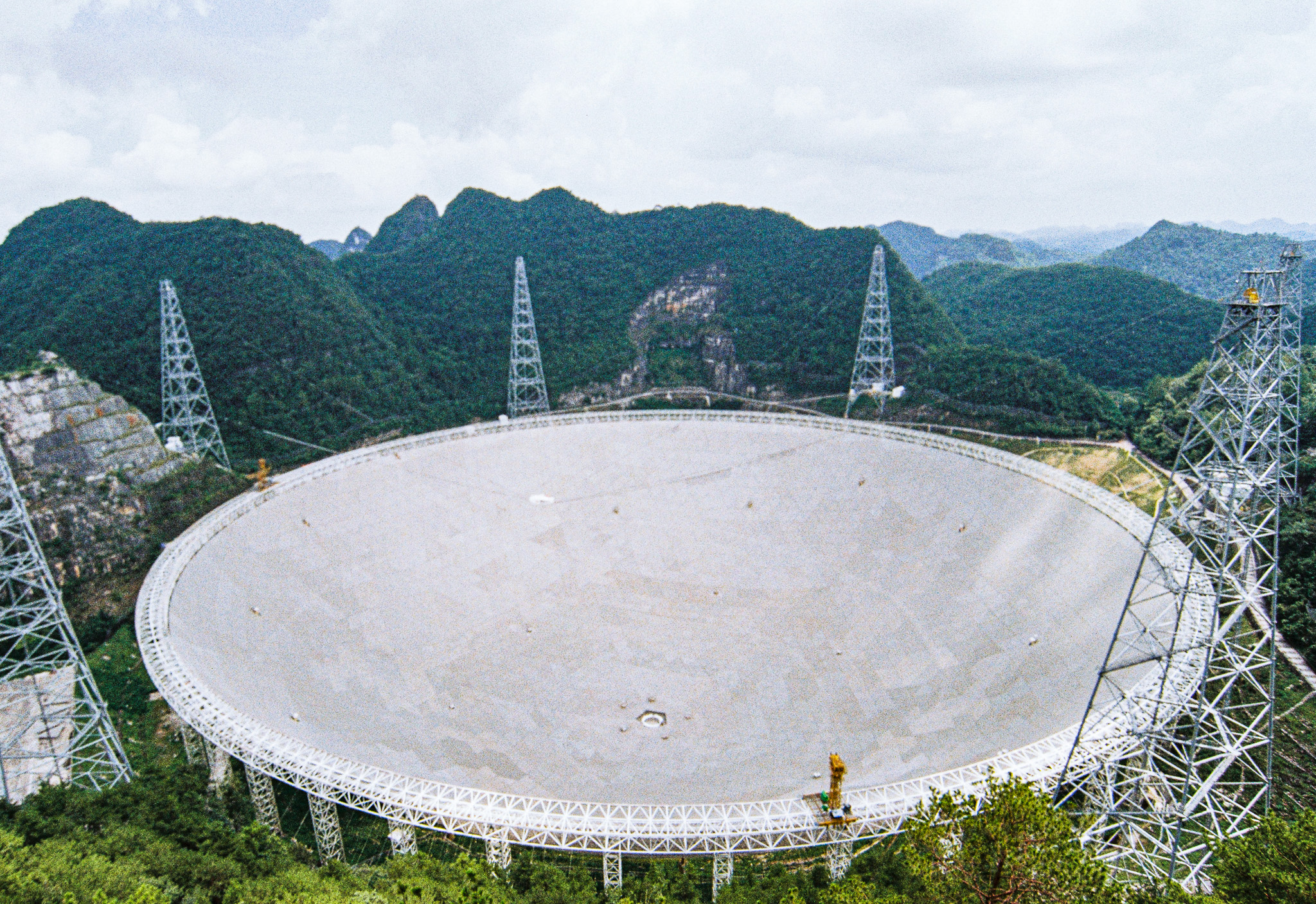 A large radio telescope dish surrounded by forested hills and several supporting towers beneath a cloudy sky, searching for signals from a distant starless gas cloud.