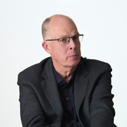 Bald man wearing glasses and a dark suit jacket sits against a plain white background, looking slightly to the side with a neutral expression.