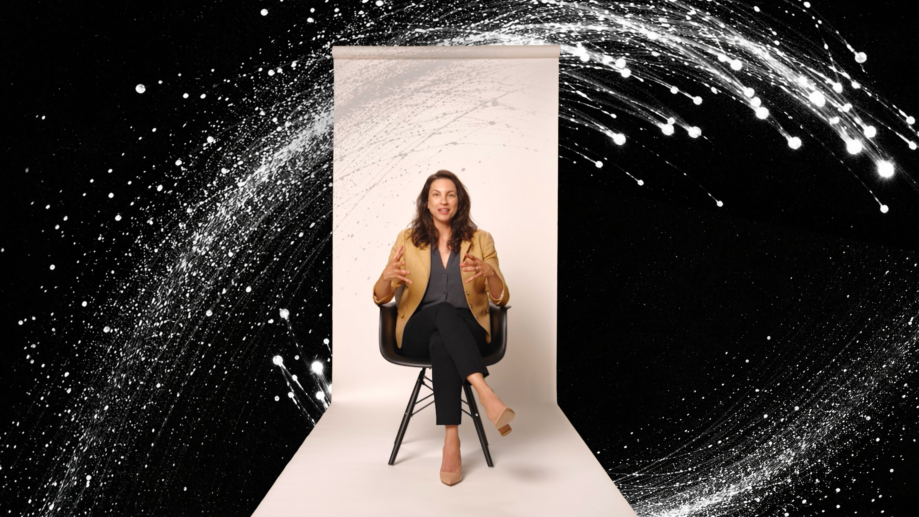 A woman sits on a chair against a white backdrop, gesturing with her hands, with a dynamic black background and white abstract swirl surrounding her.