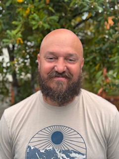 A bearded man with a shaved head is standing outside, smiling, wearing a light-colored T-shirt with a circular graphic design. Green foliage is visible in the background.