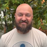 A bearded man with a shaved head is standing outside, smiling, wearing a light-colored T-shirt with a circular graphic design. Green foliage is visible in the background.