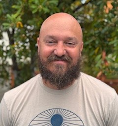 A bearded man with a shaved head is standing outside, smiling, wearing a light-colored T-shirt with a circular graphic design. Green foliage is visible in the background.