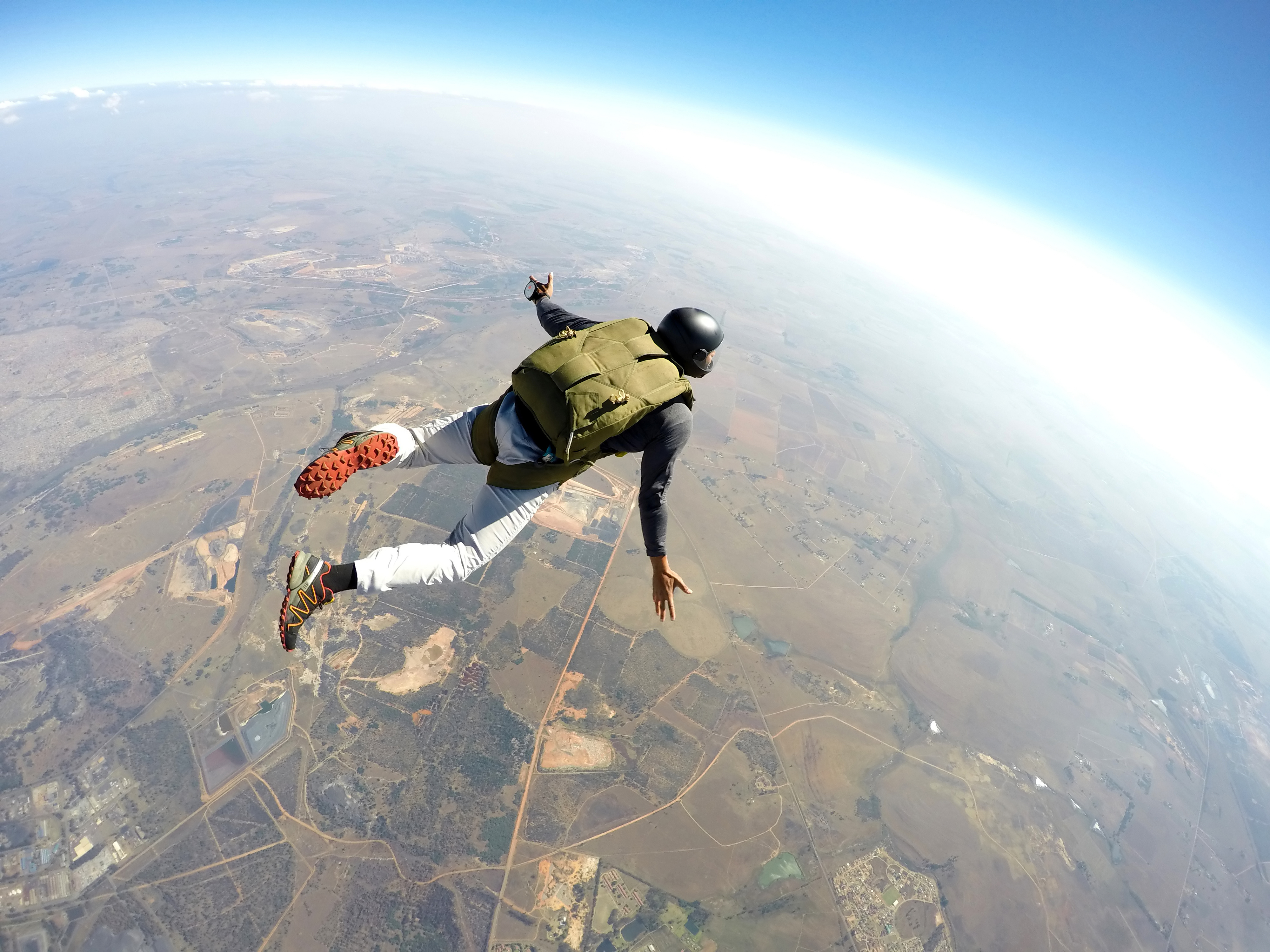 A person wearing a helmet and parachute gear is skydiving above a rural landscape with fields and roads visible below.