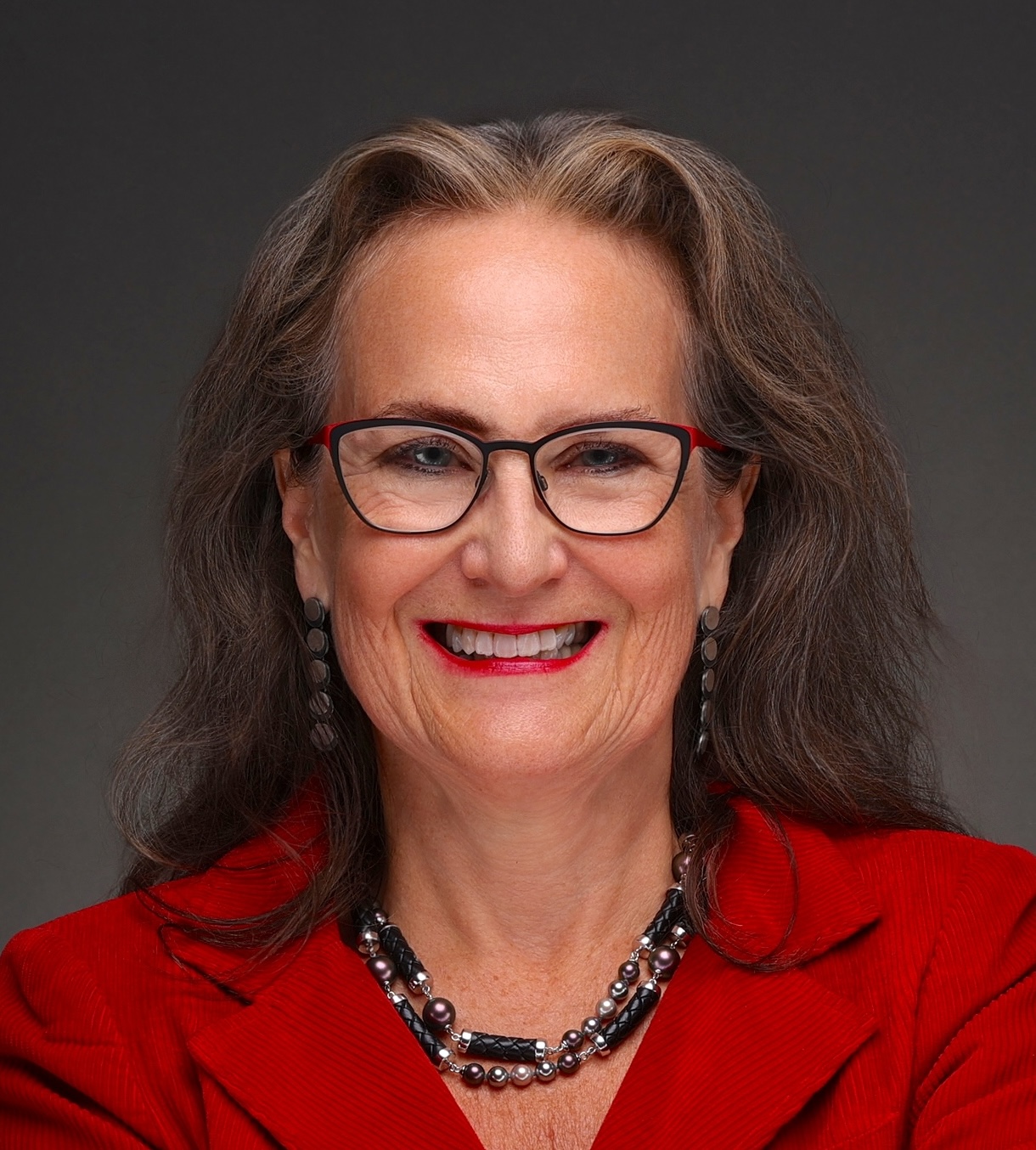 Smiling woman with long gray hair, glasses, and red lipstick wearing a red blazer, black and silver necklace, and matching earrings against a dark background.