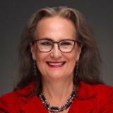 Smiling woman with long gray hair, glasses, and red lipstick wearing a red blazer, black and silver necklace, and matching earrings against a dark background.