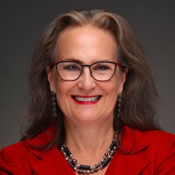 Smiling woman with long gray hair, glasses, and red lipstick wearing a red blazer, black and silver necklace, and matching earrings against a dark background.