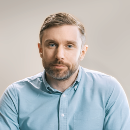 A man with short brown hair and a beard, wearing a light blue button-up shirt, sits against a plain beige background, looking directly at the camera.