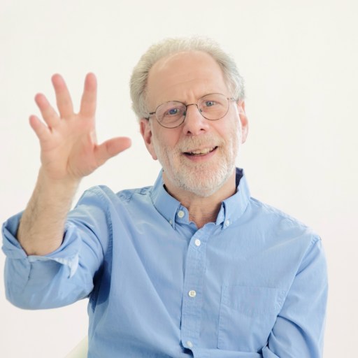 An older man with glasses and a light blue shirt sits and smiles while raising his hand with fingers spread, in front of a plain white background.