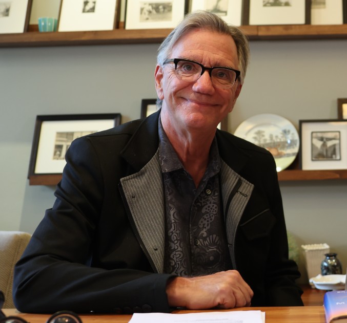 An older man with glasses and gray hair sits at a desk, smiling, with framed photos and decorative items on shelves behind him.
