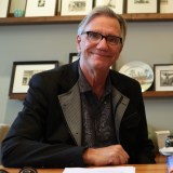 An older man with glasses and gray hair sits at a desk, smiling, with framed photos and decorative items on shelves behind him.