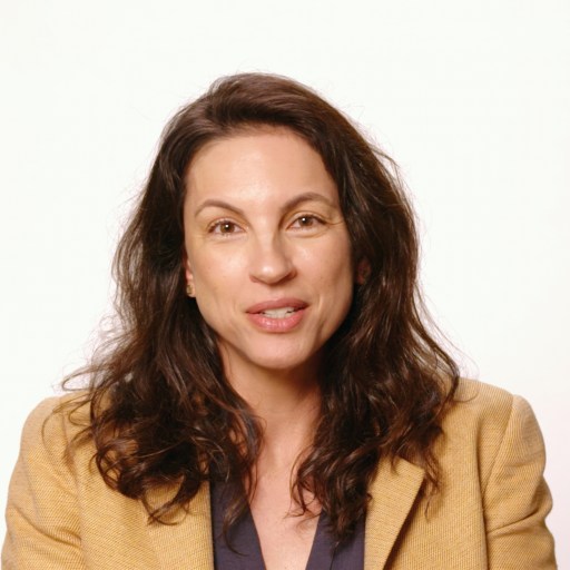 A woman with long brown hair wearing a tan blazer over a dark shirt sits in front of a plain white background, looking at the camera.