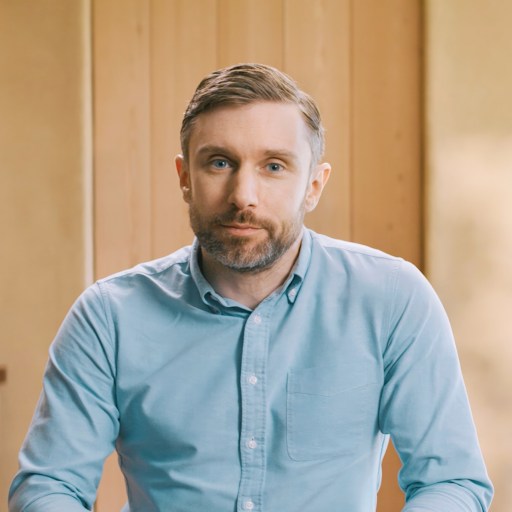 A man with short hair and a beard, wearing a light blue button-up shirt, sits indoors in front of a wooden wall.