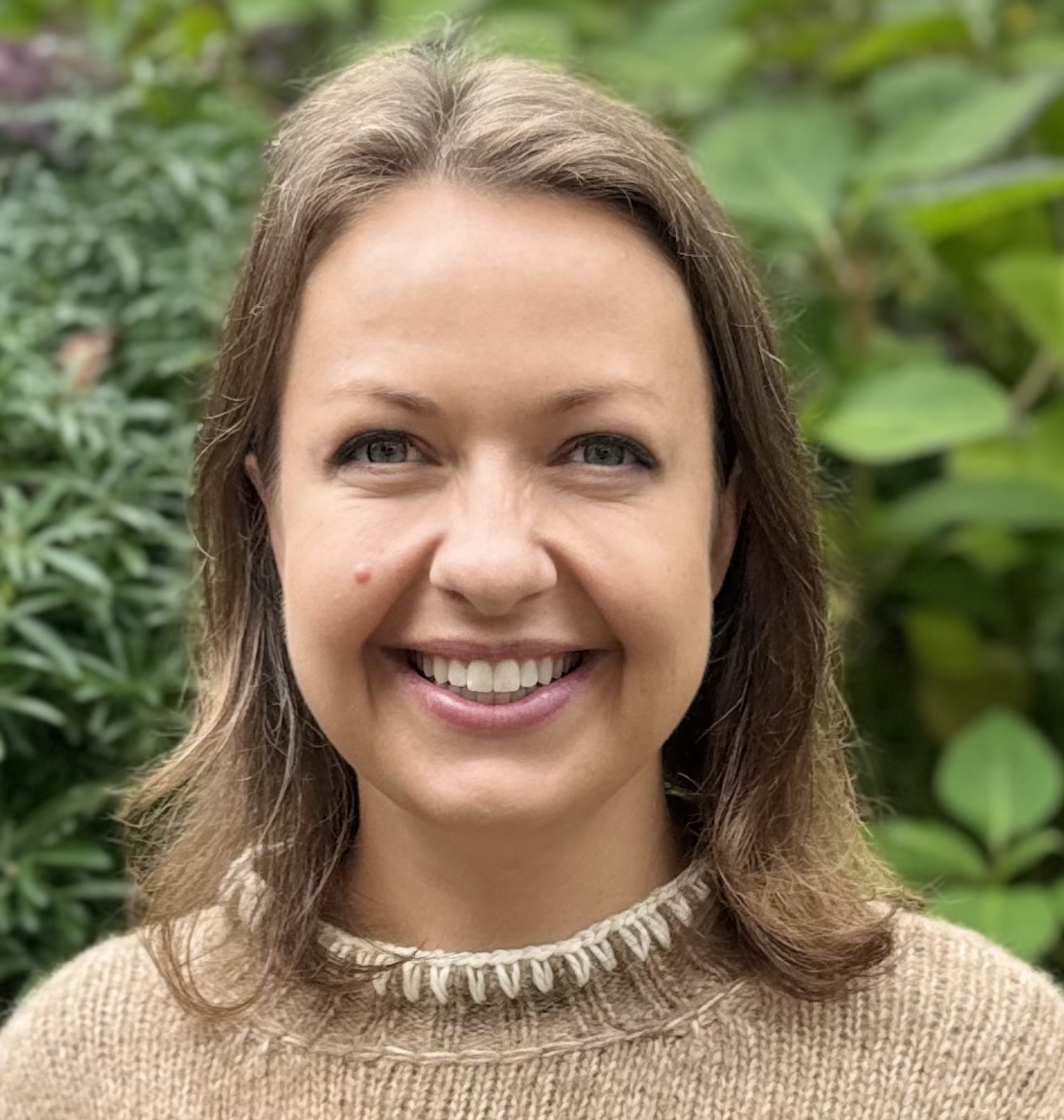 A woman with light brown hair and a beige sweater smiles at the camera with green foliage in the background.
