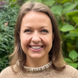 A woman with light brown hair and a beige sweater smiles at the camera with green foliage in the background.