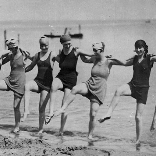Seven women in vintage swimsuits and swim caps, reminiscent of an Aaron Hurst photograph, stand in a line on the beach, kicking up one leg in unison with the sea in the background.