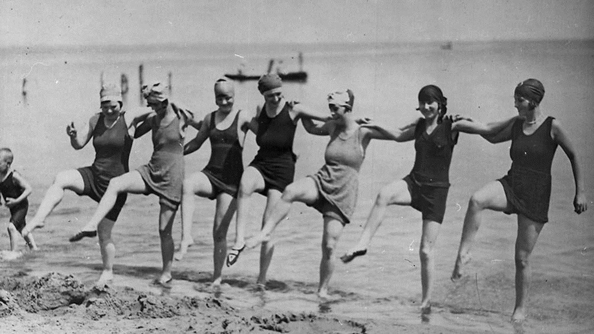 Seven women in vintage swimsuits and swim caps, reminiscent of an Aaron Hurst photograph, stand in a line on the beach, kicking up one leg in unison with the sea in the background.