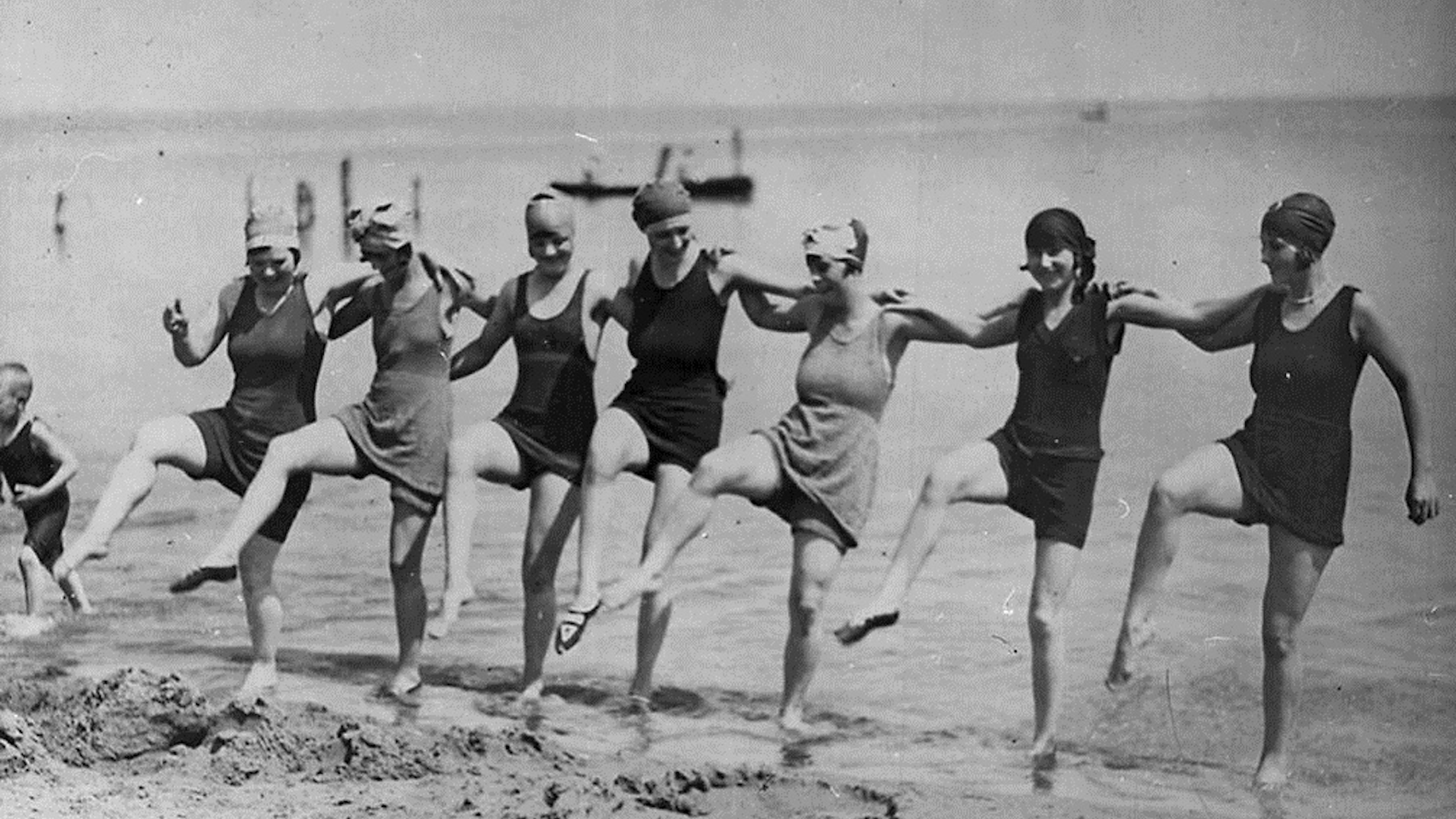 Seven women in vintage swimsuits and swim caps, reminiscent of an Aaron Hurst photograph, stand in a line on the beach, kicking up one leg in unison with the sea in the background.