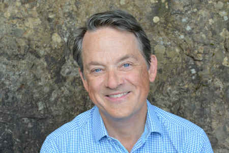 A middle-aged man with short brown hair and blue eyes smiles at the camera, wearing a light blue checkered shirt. Rock wall background.