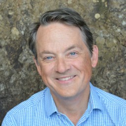 A middle-aged man with short brown hair and blue eyes smiles at the camera, wearing a light blue checkered shirt. Rock wall background.