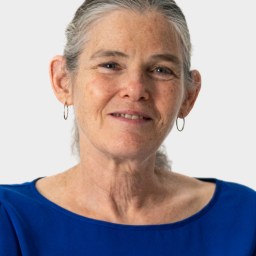 An older woman with gray hair pulled back, wearing hoop earrings and a bright blue top, is facing the camera and smiling slightly against a plain light background.