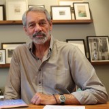 An older man with gray hair and beard sits at a desk in front of a wall with framed photos, wearing a light button-up shirt and a watch.