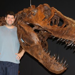 A man stands smiling next to a large dinosaur skeleton, likely a Tyrannosaurus rex, against a dark background.