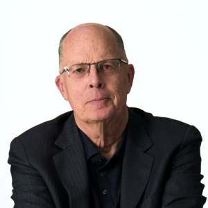 An older man with short hair, glasses, and a serious expression wears a dark suit jacket and shirt, seated against a plain white background.