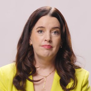 A woman with long dark hair, wearing a yellow blazer and gold necklace, looks towards the camera with a neutral expression against a plain light background.