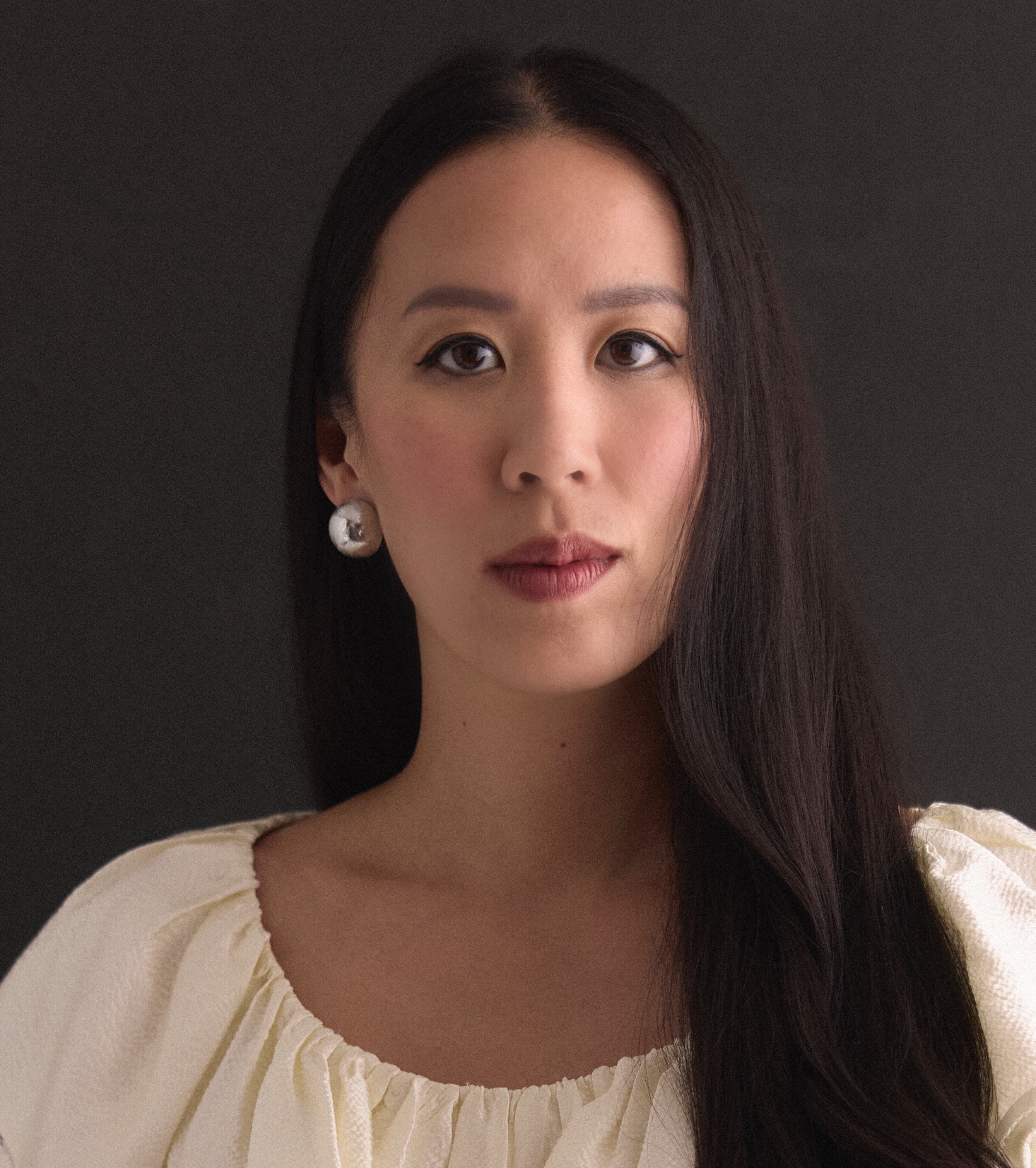 A woman with long dark hair, wearing a white blouse and round silver earrings, looks directly at the camera against a dark background.