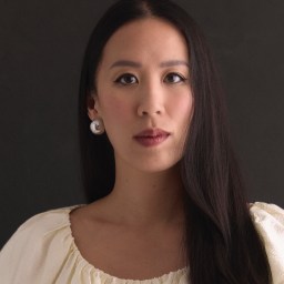 A woman with long dark hair, wearing a white blouse and round silver earrings, looks directly at the camera against a dark background.