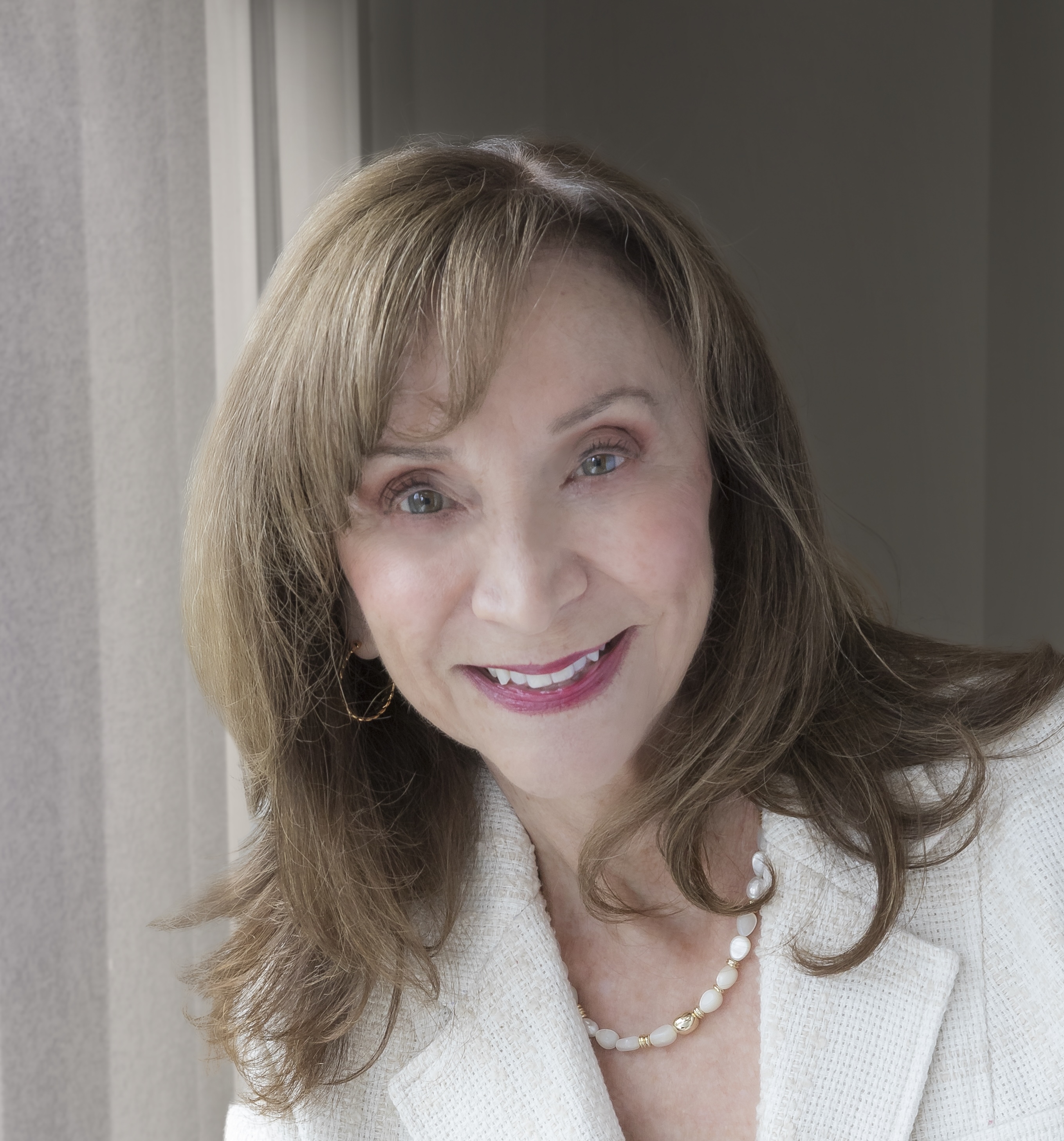 A woman with light brown hair wearing a white jacket and pearl necklace smiles near a window with natural light.