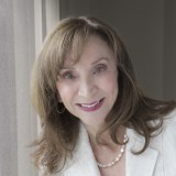 A woman with light brown hair wearing a white jacket and pearl necklace smiles near a window with natural light.