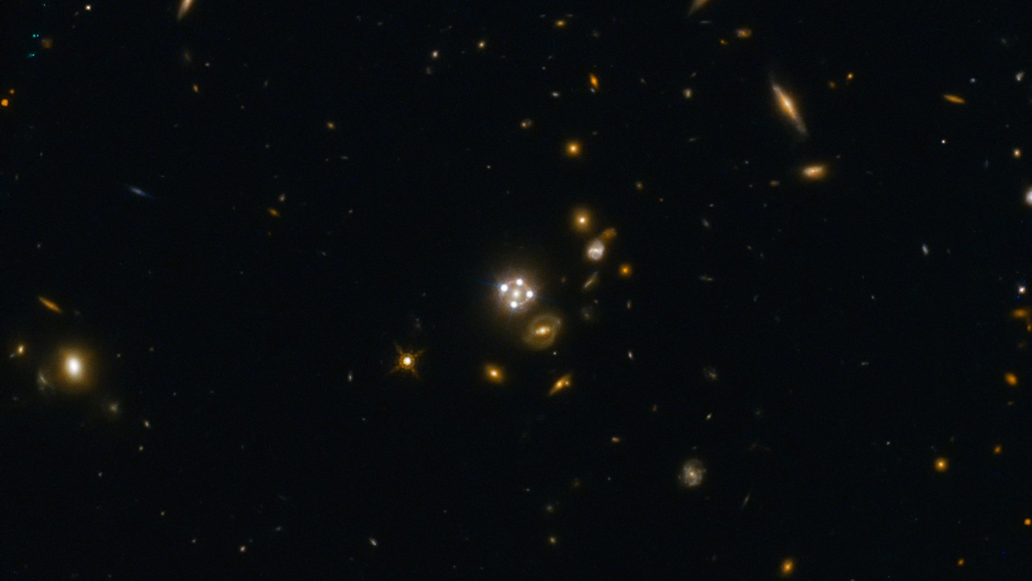 A deep space image shows numerous distant galaxies and stars against a dark background, including several bright spots shaped by a gravitational lens cross, with diffuse light sources scattered throughout.