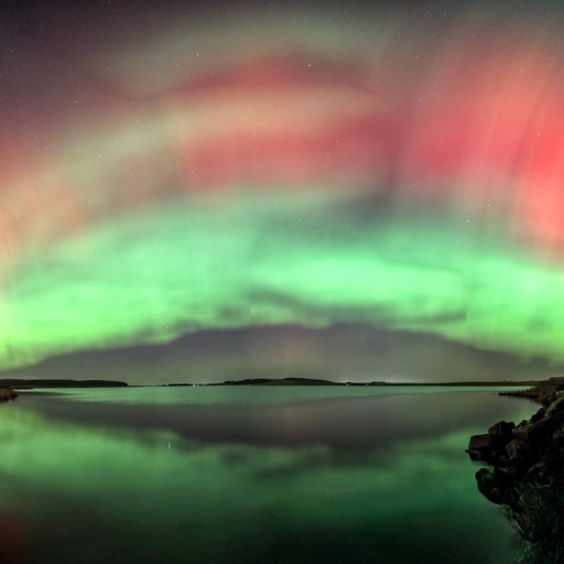 Green and red aurora borealis lights, sparked by a recent solar radiation storm, arc across the night sky and reflect over a calm lake with a rocky shoreline.