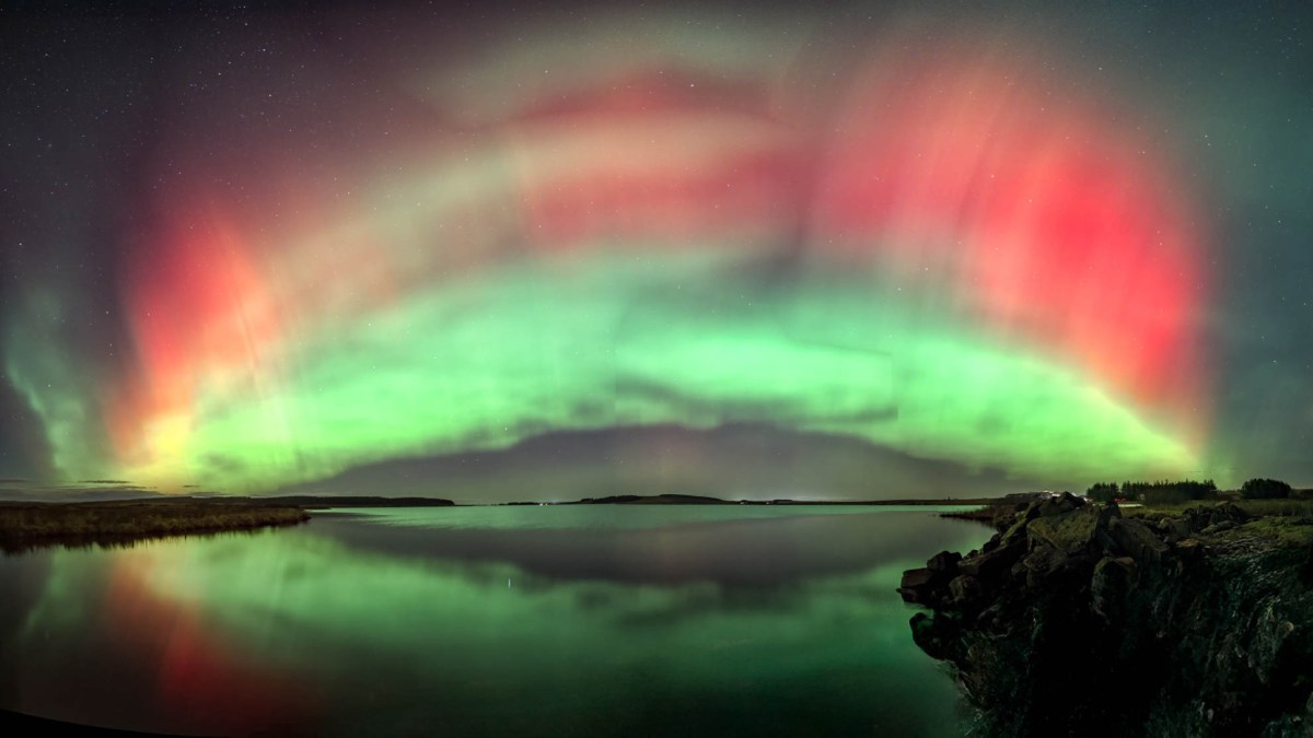 Green and red aurora borealis lights, sparked by a recent solar radiation storm, arc across the night sky and reflect over a calm lake with a rocky shoreline.