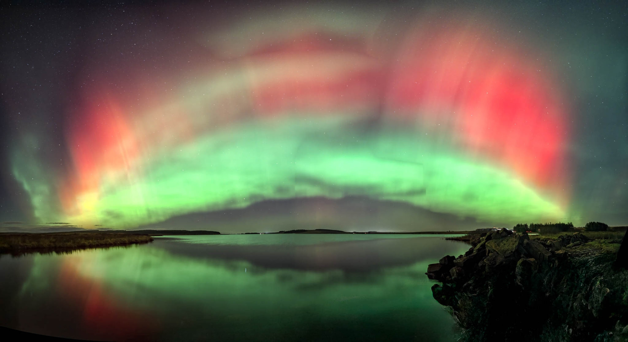 Green and red aurora borealis lights, sparked by a recent solar radiation storm, arc across the night sky and reflect over a calm lake with a rocky shoreline.