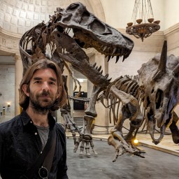 A man stands in front of a T. rex and Triceratops skeleton display in a museum with high, ornate ceilings and chandeliers.