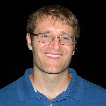 A man with short brown hair and glasses smiles at the camera. He is wearing a blue collared shirt and standing against a black background.