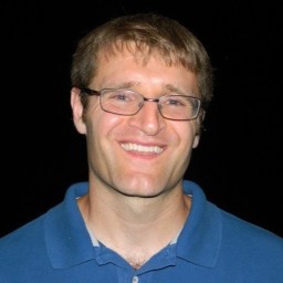 A man with short brown hair and glasses smiles at the camera. He is wearing a blue collared shirt and standing against a black background.