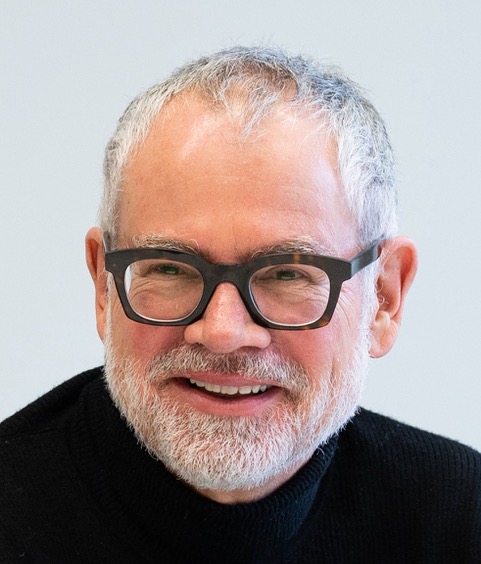 A smiling older man with short gray hair, a beard, and black glasses, wearing a black sweater against a light background.