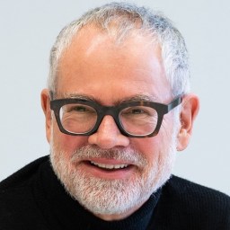 A smiling older man with short gray hair, a beard, and black glasses, wearing a black sweater against a light background.