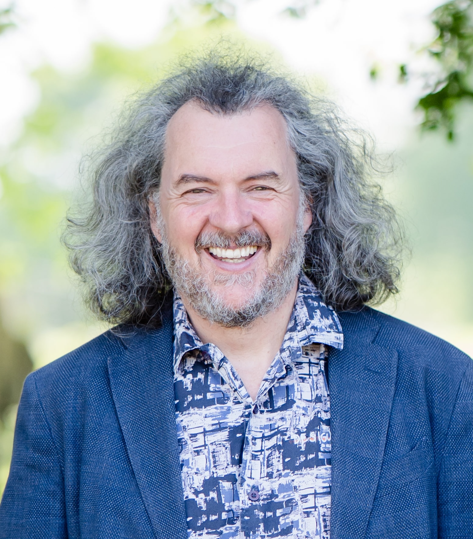 A man with gray, curly hair and a beard smiles outdoors, wearing a patterned shirt and blue blazer.