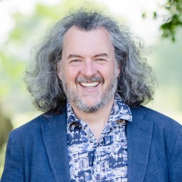 A man with gray, curly hair and a beard smiles outdoors, wearing a patterned shirt and blue blazer.