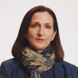 A woman with straight brown hair wears a patterned scarf and a blue jacket, posed against a plain white background.