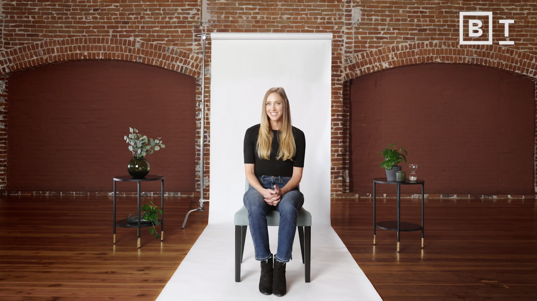 A woman sits on a chair in front of a white backdrop in a brick-walled room, with potted plants on tables on either side.