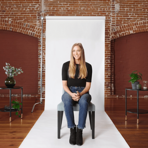 A woman sits on a chair in front of a white backdrop in a brick-walled room, with potted plants on tables on either side.