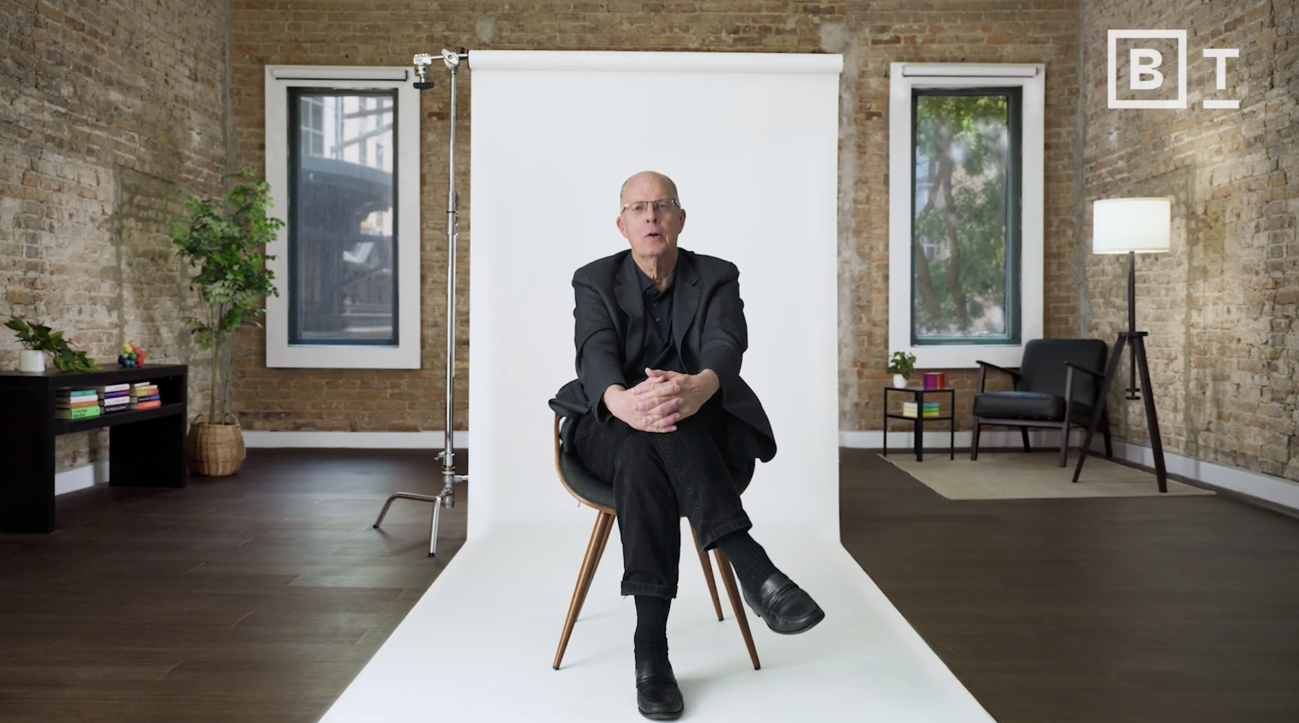 A man in a black suit sits on a chair in front of a white backdrop in a modern brick-walled room with large windows and minimal furnishings.