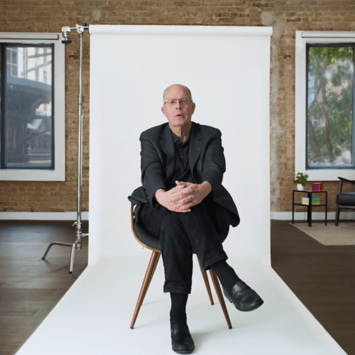 A man in a black suit sits on a chair in front of a white backdrop in a modern brick-walled room with large windows and minimal furnishings.