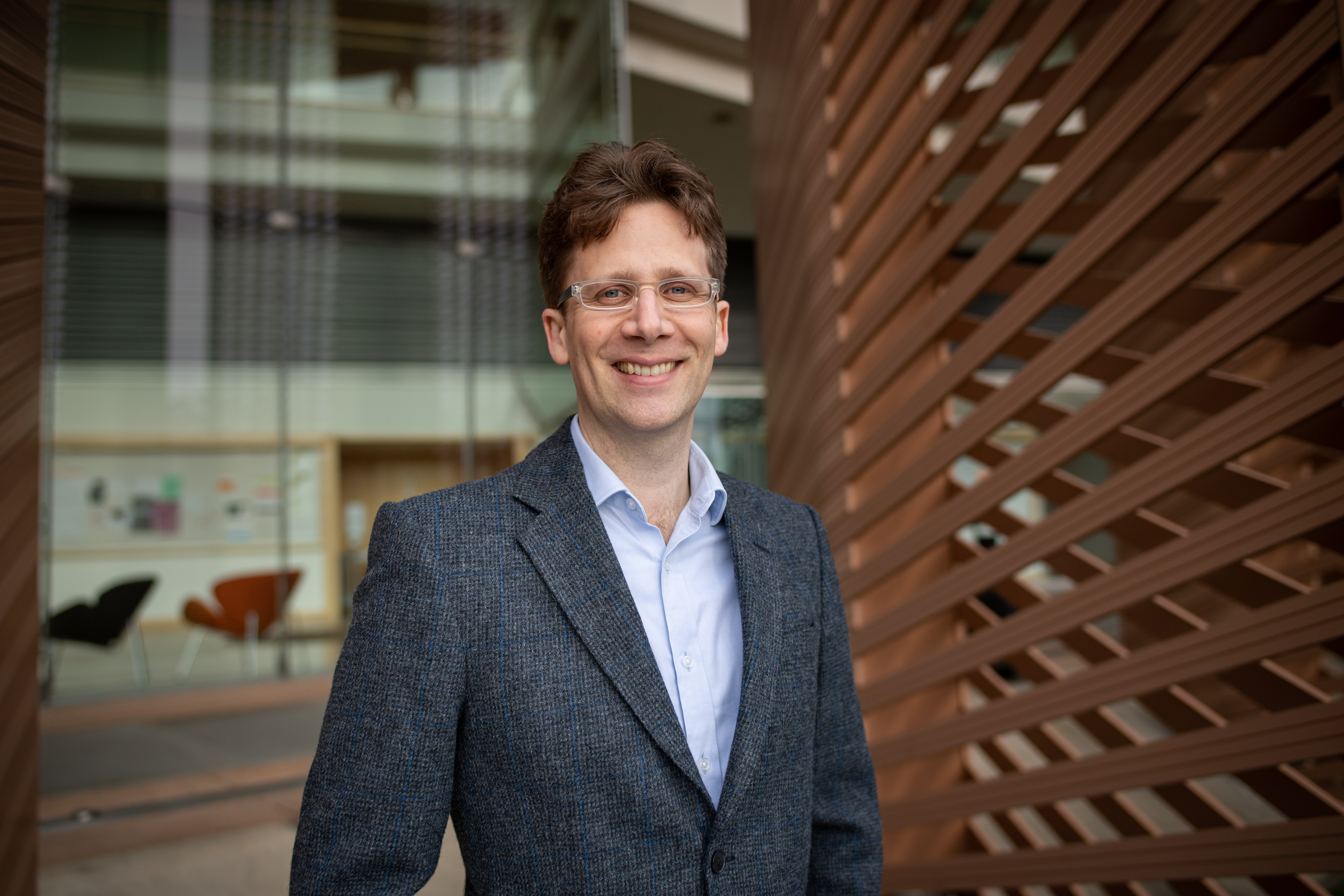 A man wearing glasses, a blue shirt, and a grey blazer stands outdoors, smiling, with a modern building and wooden slats in the background.